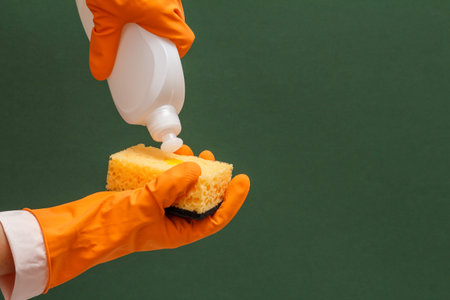 Women's hands in orange protective glove with bottle of dishwashing liquid and sponge on the green...