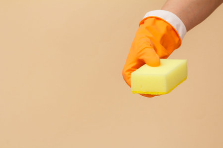 Woman's hand in orange rubber glove with sponge on beige background. Washing and cleaning concept.