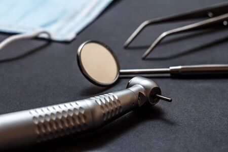 Set of metal dental instruments for dental treatment. High-speed dental handpiece, dental mirror, tweezers and mask on black background. Medical tools. Shallow depth of field. Focus on a bur.の写真素材