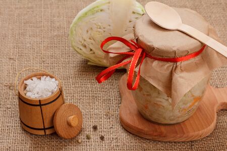 Homemade fermented cabbage with carrot in glass jar, salt in wooden small barrel and head of fresh cabbage on the sackcloth.の写真素材