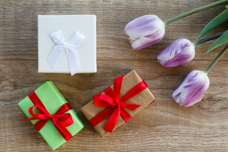 Gift boxes with red ribbons and beautiful tulips on the wooden boards. Top view. Concept of giving a...