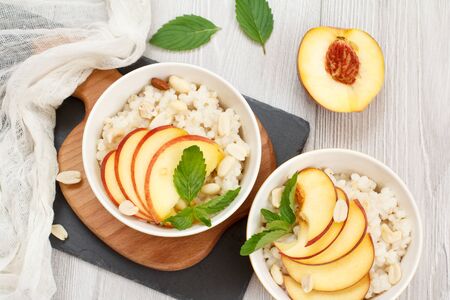 Sorghum porridge with pieces of peach, cashew nuts and almond in porcelain bowls, fresh peaches on wooden and stone boards. Vegan gluten-free sorghum salad with fruits. Top view.の写真素材