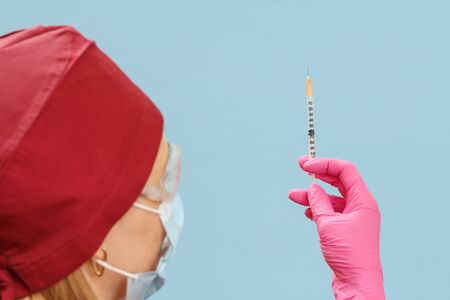Female doctor in a medical uniform, mask and latex gloves holding a syringe with liquid medicine for injection on the blueの写真素材