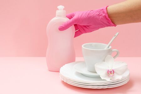 Woman's hand in a rubber protective glove with a bottle of dishwashing liquid, white plates, a saucer, a cup on the pink background. Washing and cleaning concept.の写真素材