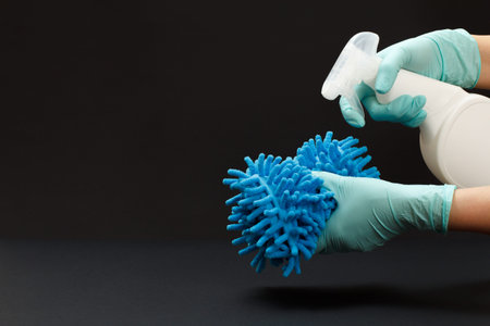 Hands in rubber gloves holding a plastic bottle of washing liquid and a rag on the black background....