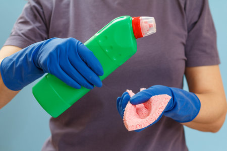Woman in a t-shirt holding a plastic bottle of washing liquid and a rag on the blue background....
