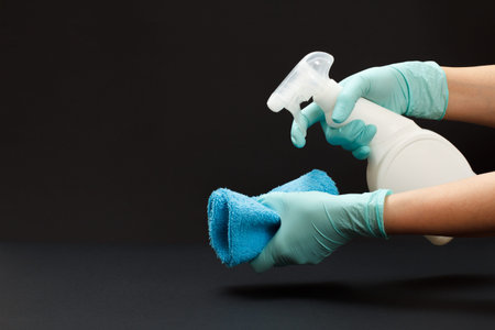 Female hands in rubber gloves holding a plastic bottle of washing liquid and a rag on the black...