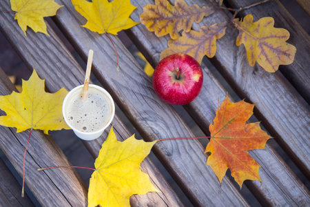 Cup of coffee, an apple and yellow dry leaves on a wooden bench in the city park. Top view. Autumn...