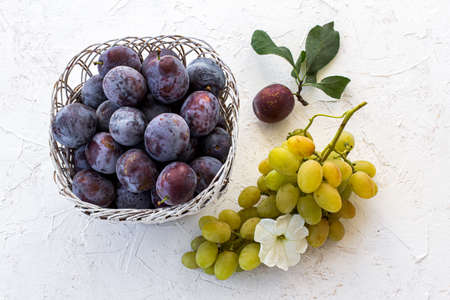 Just picked ripe plums in wicker basket and a bunch of ripe white grapes on the white structured background. Just harvested fruits. Topv view.の写真素材