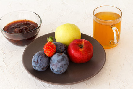 Picked ripe plums, apples, a strawberry on the plate and a glass bowl with jam, a cup of tea on the...