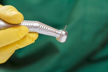 Dentist's hand in a yellow latex glove with a new high-speed dental handpiece and a green medical robe on the background. Medical tools concept.の写真素材