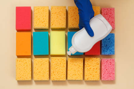 Sponges of different colors and a hand in rubber glove with plastic bottle of dishwashing liquid on the beige background. Top view. Washing and cleaning set.の写真素材