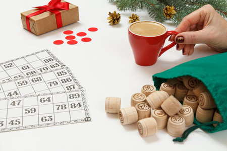 Woman's hand holding a cup of coffee. Wooden lotto barrels with a green bag and game cards. Gift box and christmas fir tree branches on the background. Board game lottoの写真素材