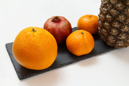 Pomegranate, an orange, tangerines and a pineapple on the black stone board and white background. Organic fruits.の写真素材