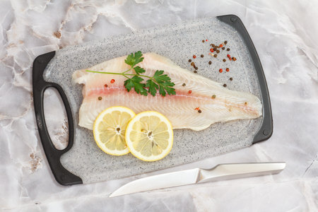 Fillet of raw pangasius fish with parsley leaves, spices and lemon on cutting board. Top view.