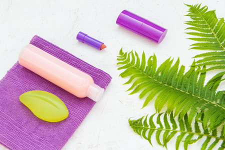 Bottle of shampoo and soap on a towel, a deodorant, a lipstick and fern leaves on the structured...
