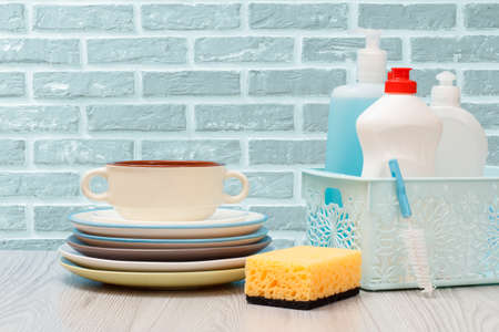 Plastic bottles of dishwashing liquid, glass and tile cleaner in basket, clean plates and a bowl on the brick wall background. Washing and cleaning concept.の写真素材