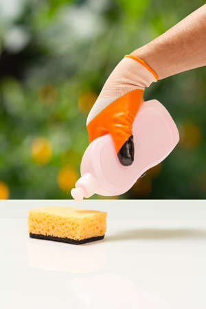 Man's hand in an orange protective glove with bottle of dishwashing liquid and a sponge. Washing and cleaning concept.の写真素材
