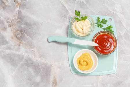 Porcelain bowls with ketchup, mustard and cheese sauce with leaves of parsley and a knife on the kitchen table. Top view.の写真素材