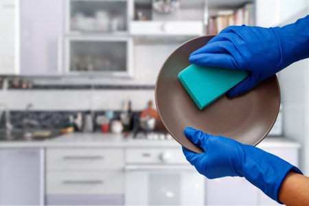 Women's hands in protective gloves with brown plate and blue sponge at the kitchen. Washing and...