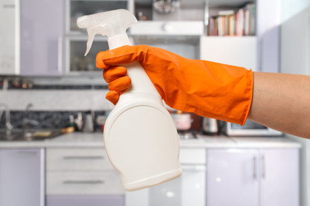 Woman's hand in orange protective glove with the bottle of glass cleaning liquid and the kitchen on...