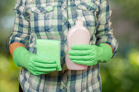Women's hands in rubber gloves holding the plastic bottle of washing liquid and the rag on the blurred natural background. Washing and cleaning concept.の写真素材