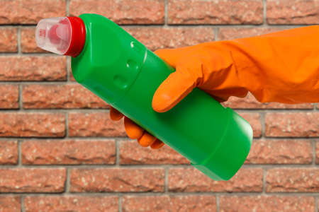 Woman's hand in orange protective glove with the bottle of washing liquid and a brick wall on the background. Washing and cleaning concept.の写真素材