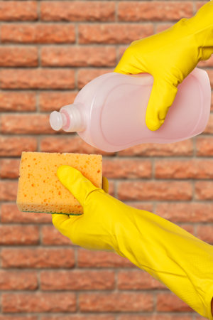 Women's hands in yellow protective gloves with a bottle of dishwashing liquid and a sponge with a...
