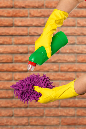 Women's hands in yellow protective gloves with a bottle of washing liquid and a rag with a brick...