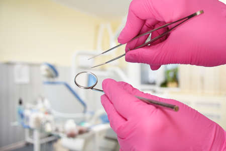 Dentist's hands in pink latex gloves with a tweezers and a mouth mirror with a dental clinic on the background. Medical tools concept.の写真素材
