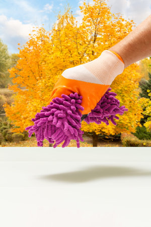 Hand in a rubber glove holding a rag with blurred autumn trees on the background. Washing and...