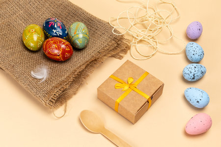 Colored Easter eggs on a sackcloth bag, a rope, a gift box and a wooden spoon on the beige...