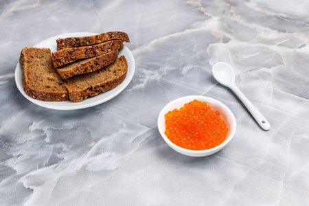Red salmon caviar in a white porcelain bowl and a plate with rye bread on the kitchen table. Top view.の写真素材