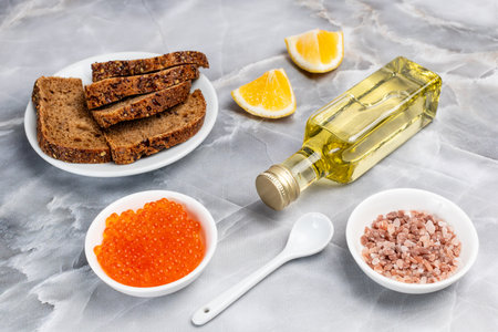 White porcelain bowls of red salmon caviar and salt, sliced lemon, a plate with rye bread and a bottle of cooking oil on the kitchen table. Top view.の写真素材