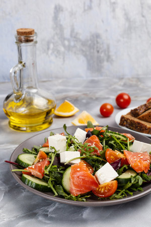 Salad with sliced salmon fish, white cheese feta and fresh vegetables on the plate. A plate with rye bread, a bottle of oil, fresh tomatoes and sliced lemon on the kitchen table.の写真素材