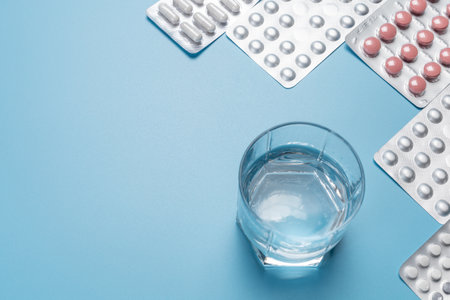 Glass of potable water and full medicine blister packs with capsules and pills on the blue background. Top view with a copy space. Concept of using medicines for treatment of diseases.の写真素材