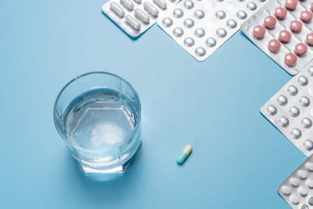 Glass of potable water and full medicine blister packs with capsules and pills on the blue background. Top view. Concept of using medicines for the prevention and treatment of diseases.の写真素材