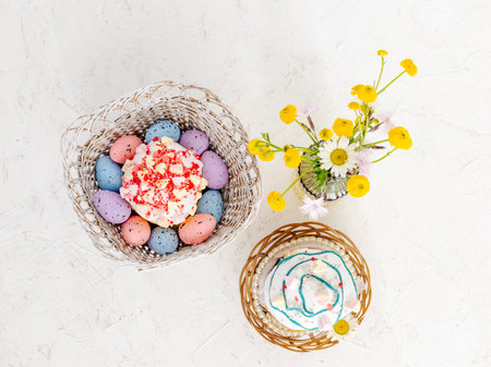 Sweet Easter cakes, colorful Easter eggs in the wooden baskets and yellow Ranunculus asiaticus in the glass vase on the white background. Top view.の写真素材