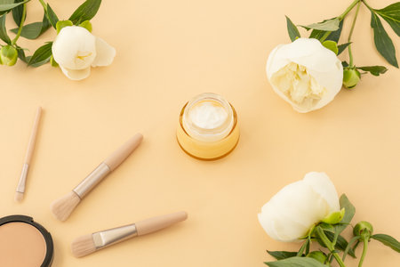 Box with powder, brushes, face cream and peony flowers on the beige background. Women cosmetics. Top view.の写真素材