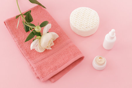 Products for facial and body care. Soft terry towel with a peony flower, a face cream, a facial serum and a sponge on the pink background. Spa and body care set. Top view.の写真素材