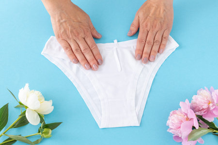 Women's hands holding a bikini-brief style cotton panties on the blue background with peony flowers....