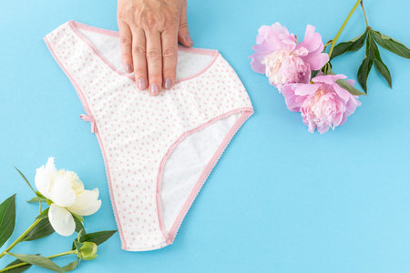 Woman's hand holding a bikini-brief style cotton panties on the blue background with peony flowers....