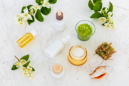 Products for facial and body care. Bottles of facial serum, face cream, a soap, aroma oil, a bowl with sea salt, a sea shell and branches of jasmine on the white background. Spa and body care set. Top view.の写真素材