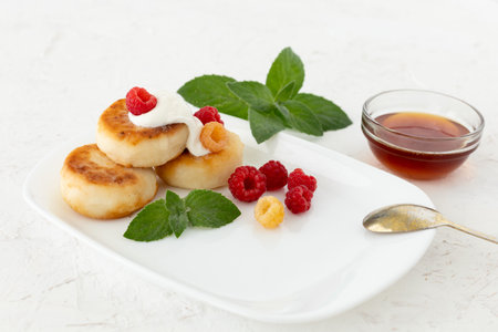 Homemade curd fritters with sour cream, raspberries and mint leaves. Glass bowl with honey on the white background.の写真素材