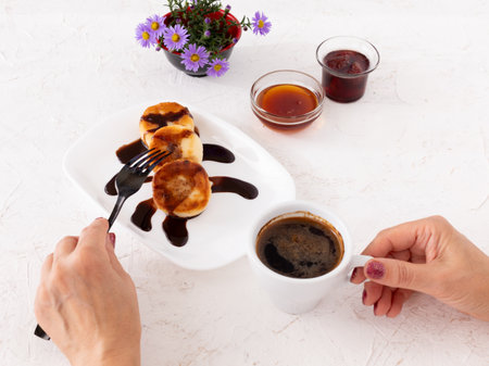 Woman's hands with a cup of coffee, a porcelain plate with homemade curd fritters decorated by chocolate, glass bowls with honey and strawberry jam, a vase with flowers. Top view.の写真素材
