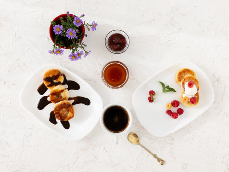 Porcelain plates with homemade curd fritters with sour cream, raspberries, chocolate. Glass bowls with strawberry jam and honey. A vase with flowers. A cup of coffee. Top view.の写真素材