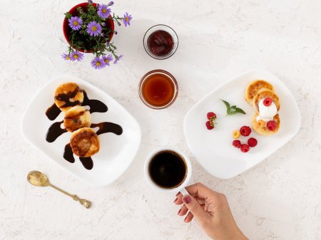 Porcelain plates with homemade curd fritters with sour cream, raspberries, chocolate. Glass bowls with strawberry jam and honey. A vase with flowers. A woman's hand with a cup of coffee. Top view.の写真素材