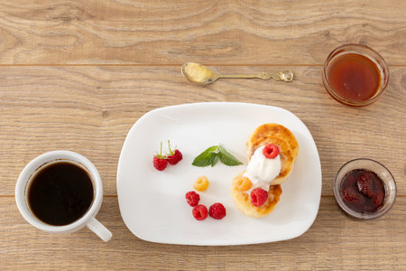 Porcelain plate with homemade curd fritters with sour cream and raspberries. Glass bowls with strawberry jam and honey, a spoon and a cup of coffee on the wooden background. Top view.の写真素材