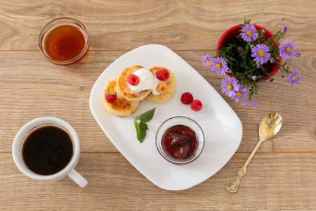 Porcelain plate with homemade curd fritters with sour cream and raspberries. Glass bowls with strawberry jam and honey, a spoon, a vase with flowers and a cup of coffee on the wooden background. Top view.の写真素材