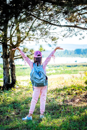 A young girl with a backpack and raised up with her hands rejoices at the sunny day. Active rest on natureの写真素材